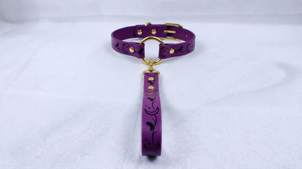A purple collar with a gold buckle and a black floral pattern on the strap is displayed against a white background.