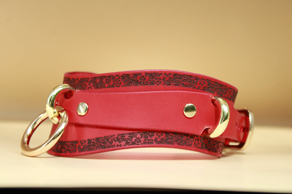 A red leather choker with a floral pattern and two gold-colored buckles is displayed on a light-colored surface.