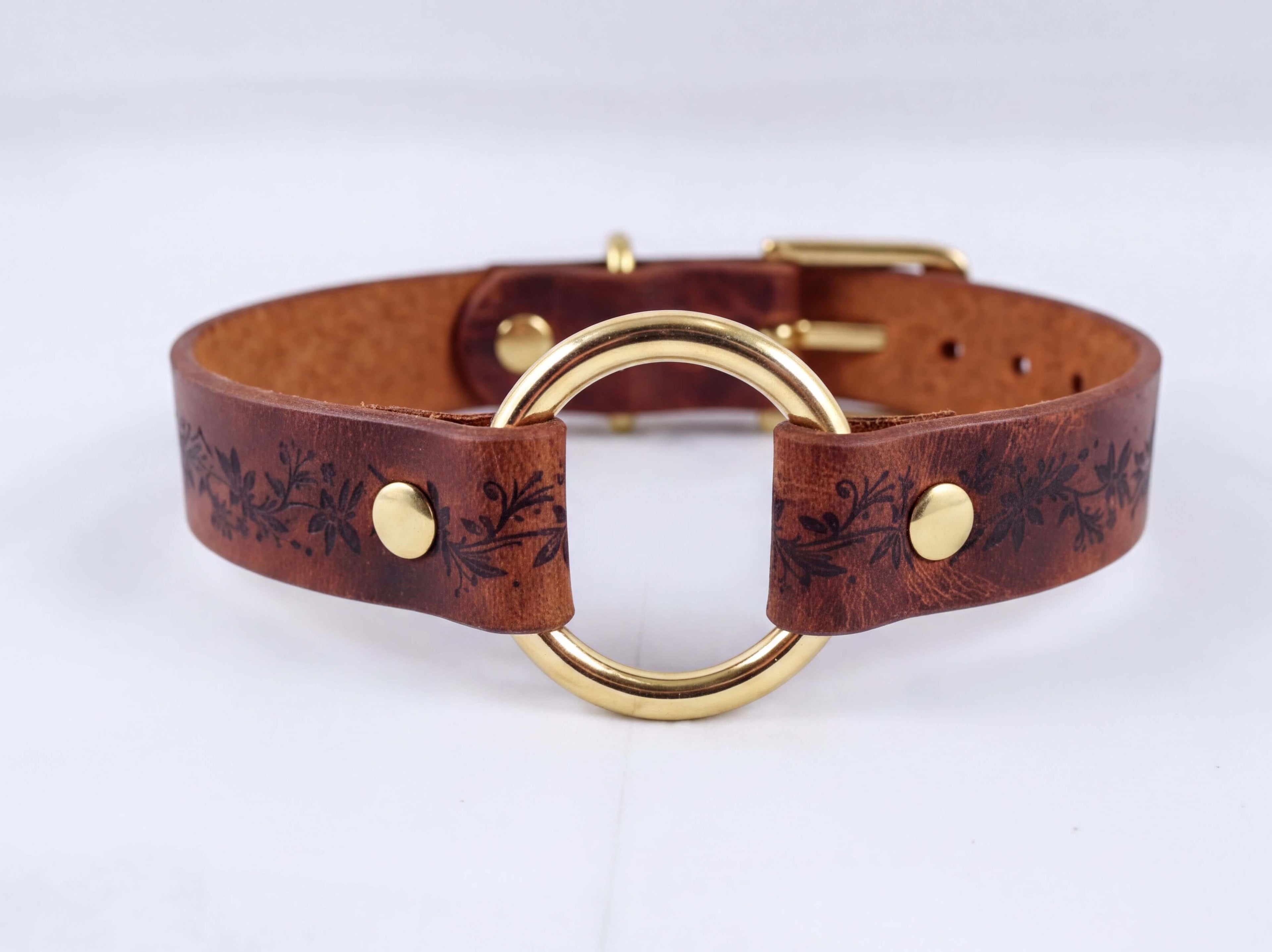 A brown leather choker collar with a gold-colored metal ring and decorative floral patterns.