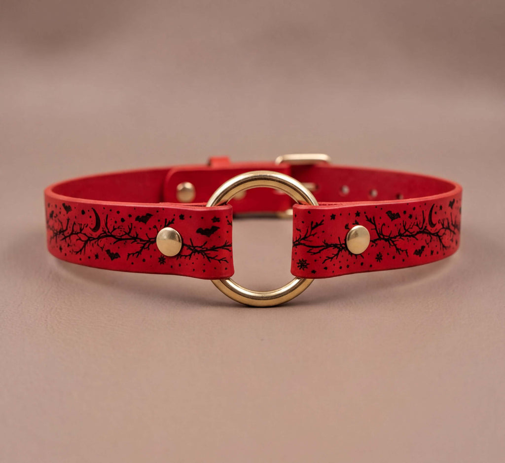 A red leather collar with a gold-colored circular buckle and a black design featuring bats and stars.