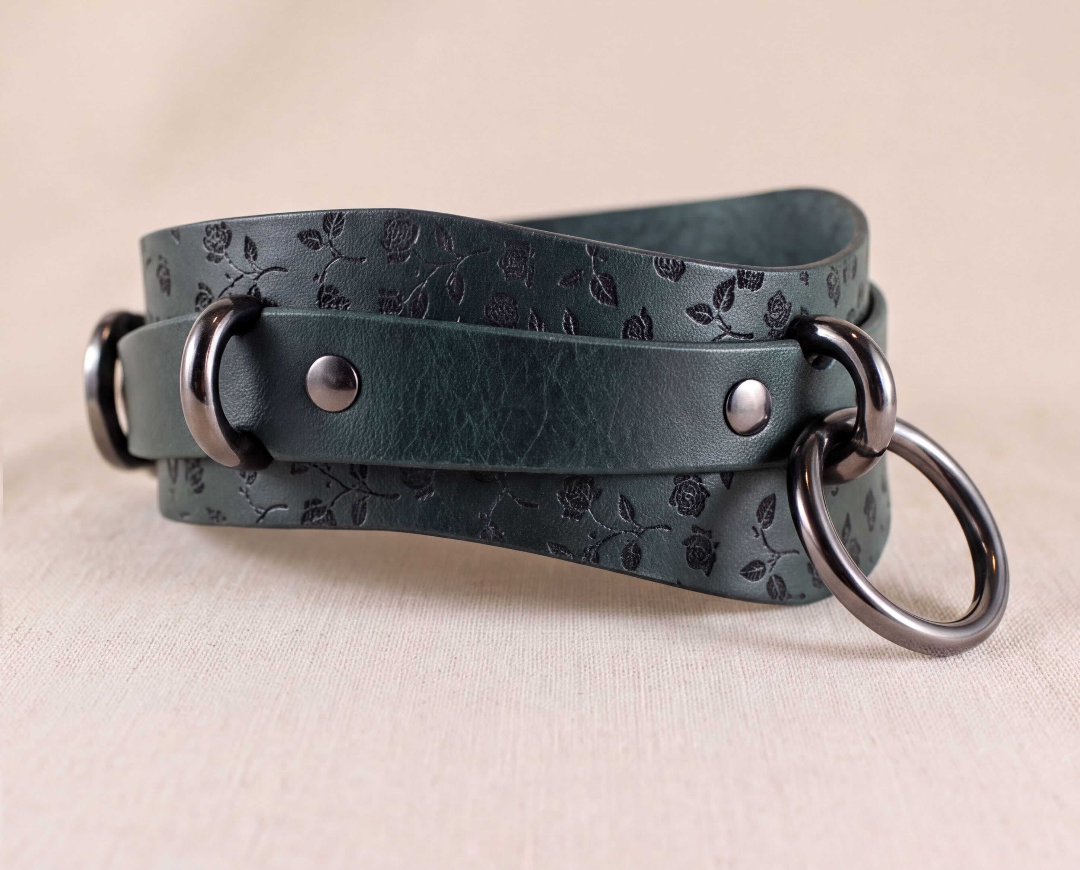 A dark green leather strap with a floral pattern and metal rings on the ends, resting on a beige background.