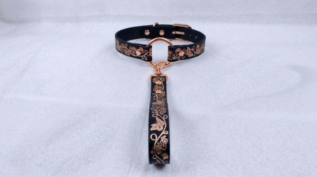A black collar with a gold-colored metal clasp and a decorative metal handle attached to it, resting on a white surface.