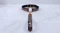 A black collar with a gold-colored metal clasp and a decorative metal handle attached to it, resting on a white surface.