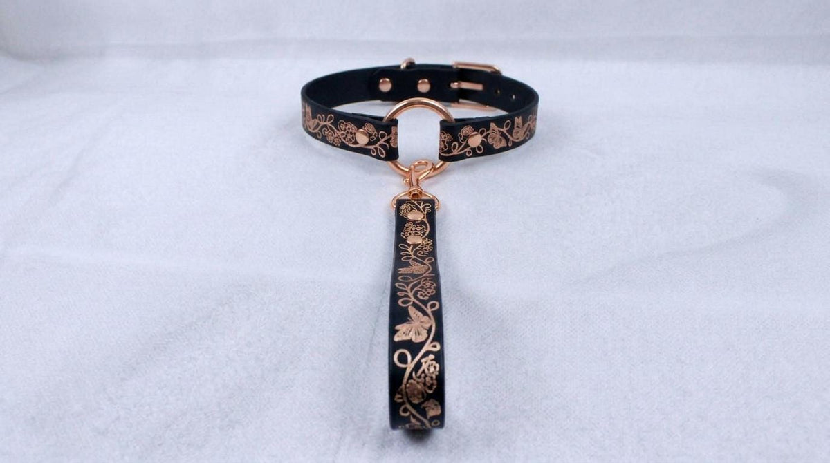 A black collar with a gold-colored metal clasp and a decorative metal handle attached to it, resting on a white surface.