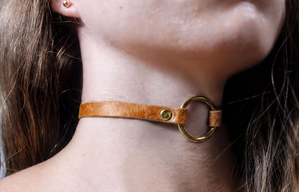 A close-up view of a person's neck and chin area, featuring a leather choker necklace with a gold-colored metal ring.