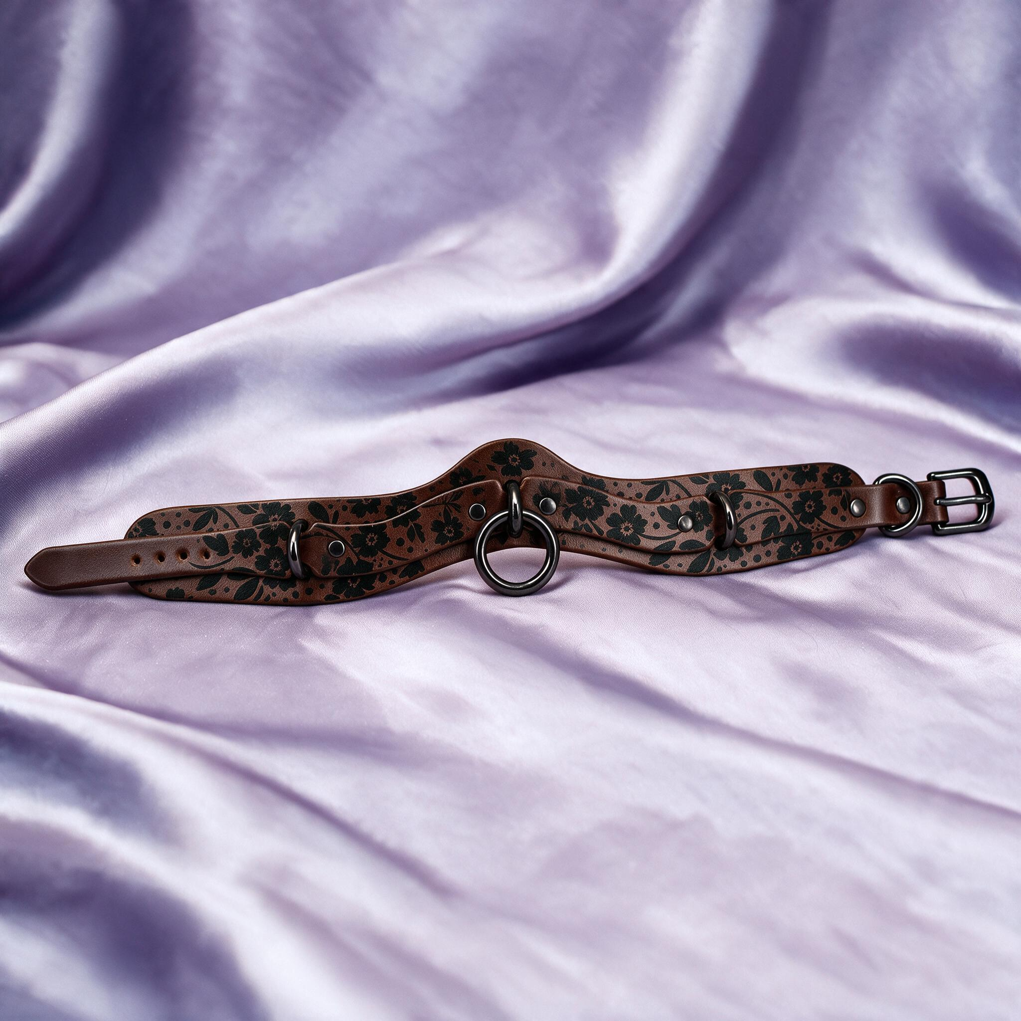 A brown leather choker necklace with a floral pattern and metal rings is displayed on a purple fabric background.
