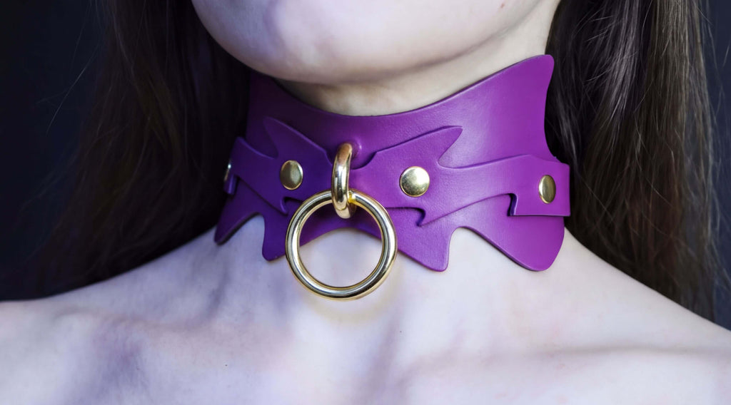 A close-up of a person's neck wearing a purple leather choker necklace with a gold ring and two gold studs on the collar.