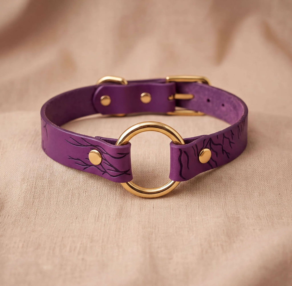 A purple leather choker collar with a gold ring and buckle, displayed on a beige fabric background.