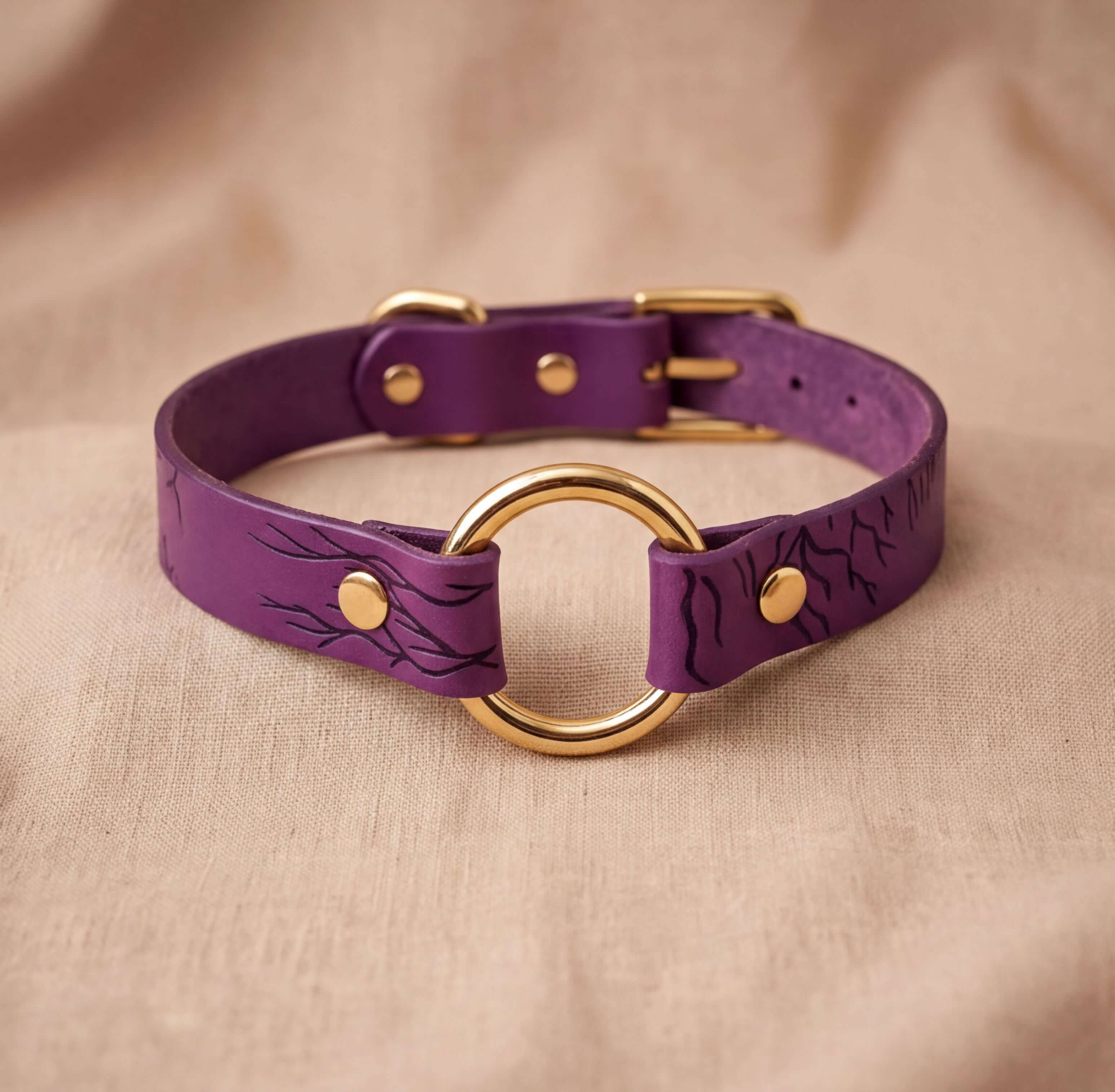 A purple leather choker collar with a gold ring and buckle, displayed on a beige fabric background.