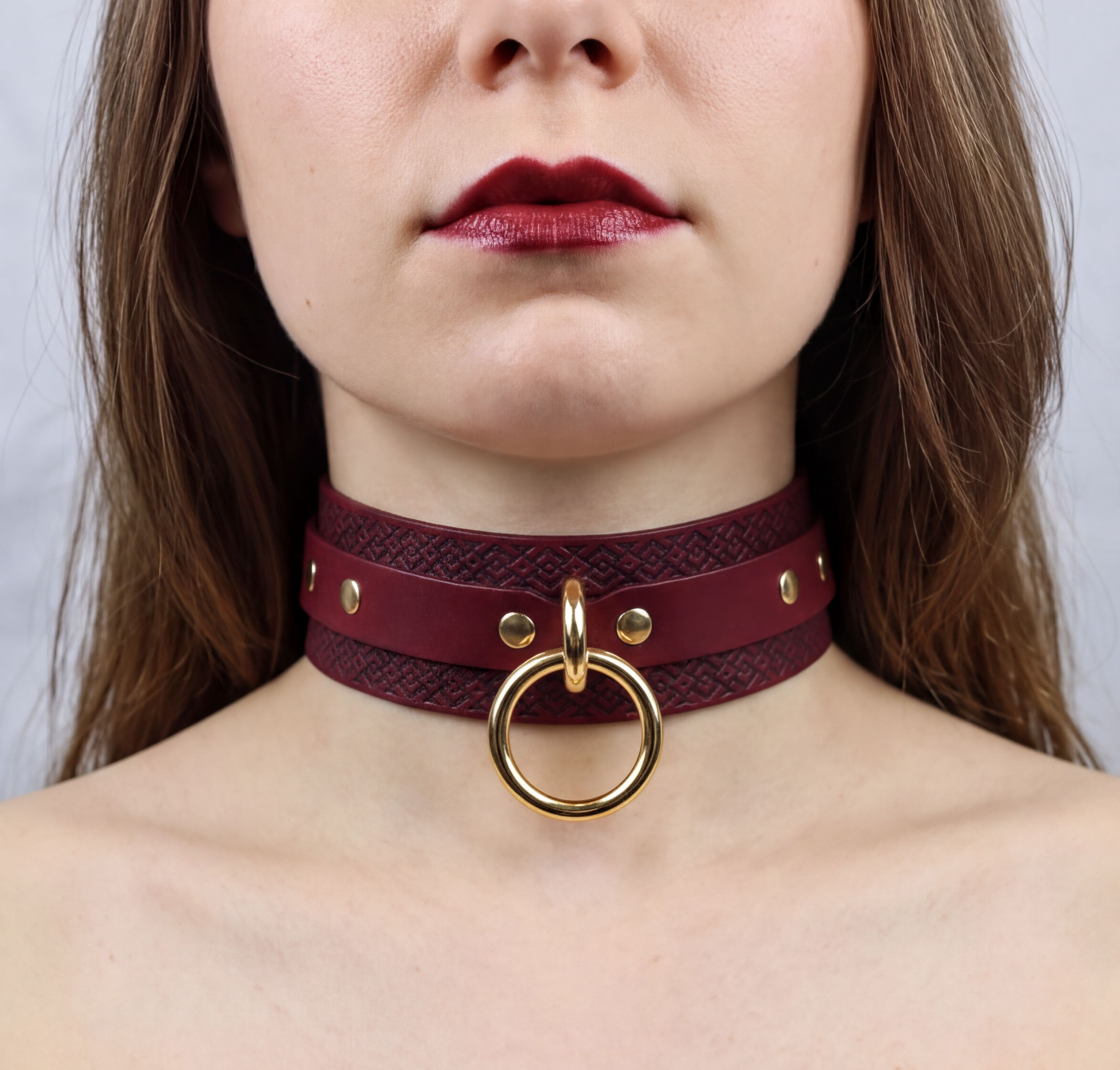 A close-up of a woman's neck and chest area, featuring a burgundy leather choker necklace with a gold ring and studs.