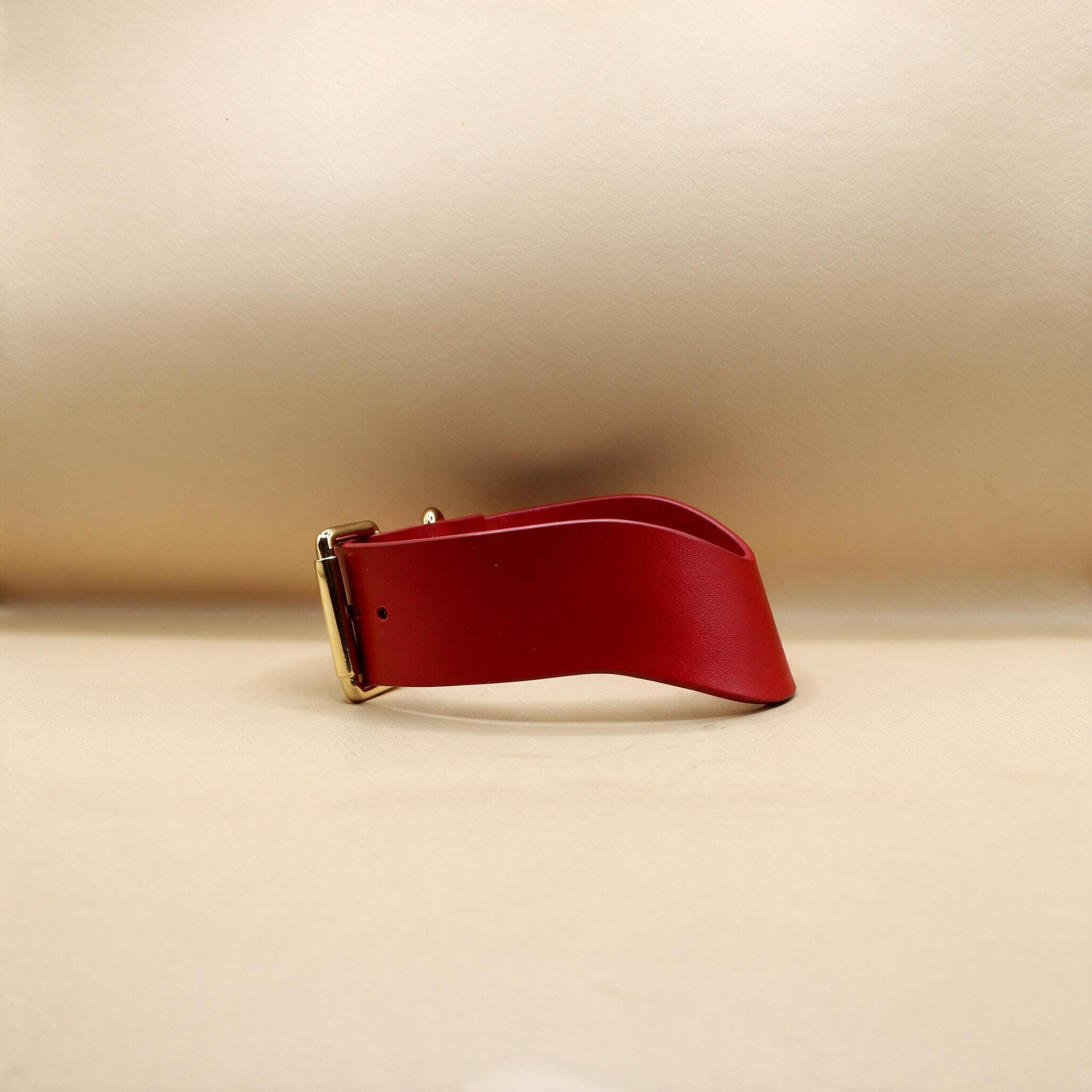 A red leather belt with a gold buckle is shown against a beige background.