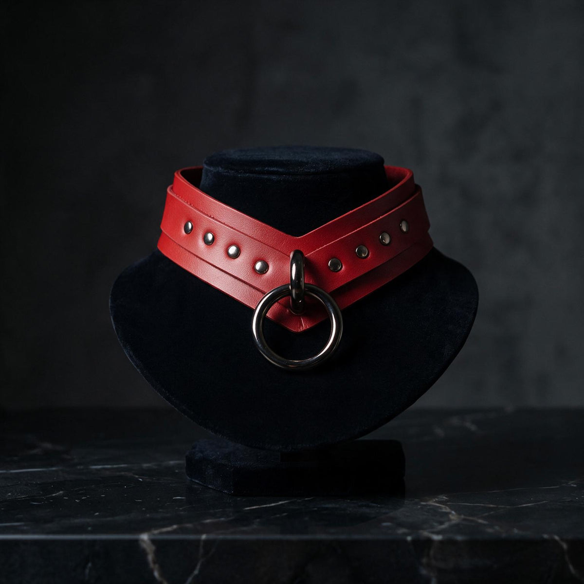 A red leather choker necklace with a large circular metal ring is displayed on a black velvet stand against a dark background.