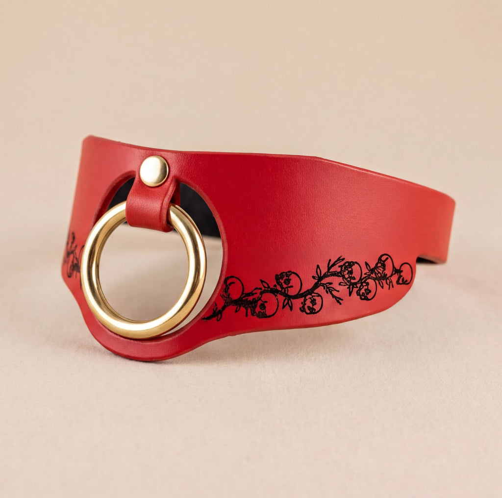 A red leather choker necklace with a gold-colored ring and a black floral design on the side.