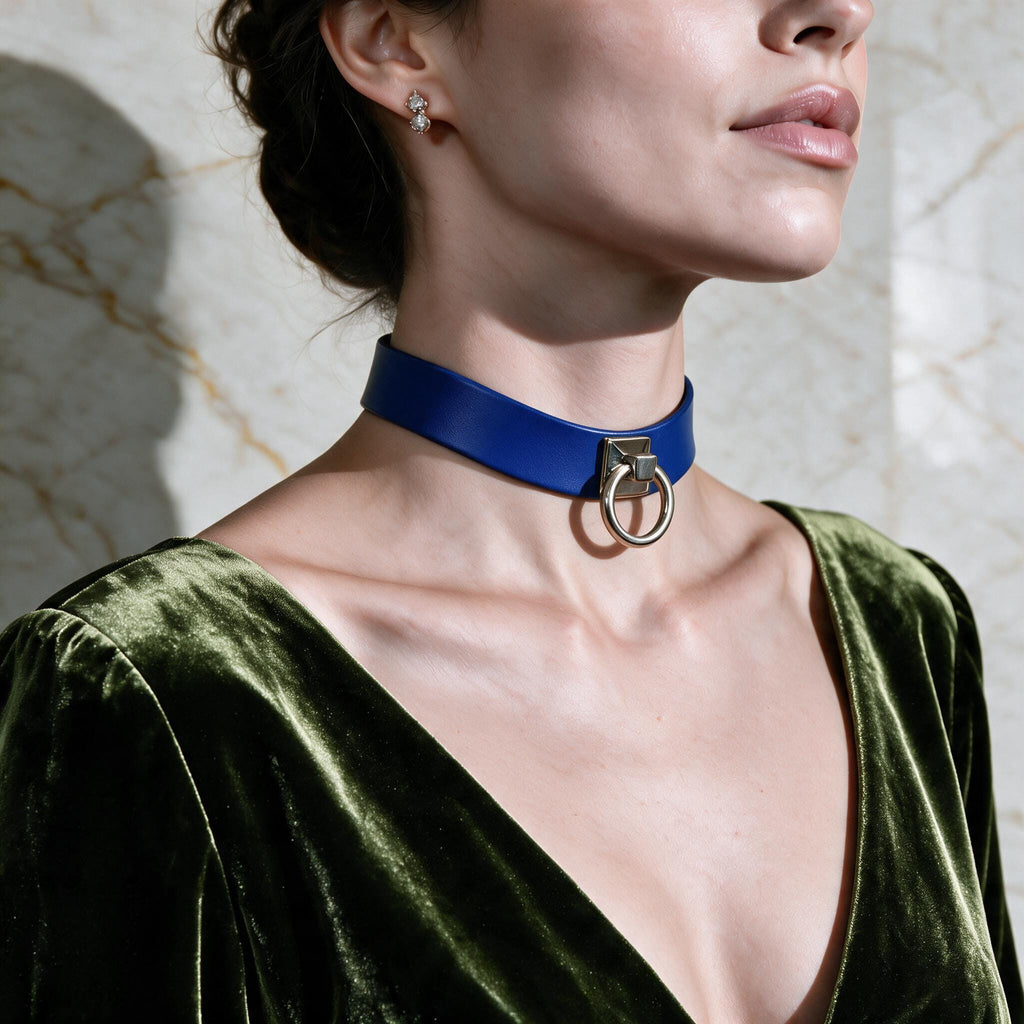 A woman wearing a blue choker necklace with a metal ring on it, standing against a marble wall.