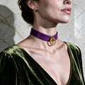 A woman wearing a purple choker necklace with a gold ring on it, and a green velvet dress, posing against a marble wall.