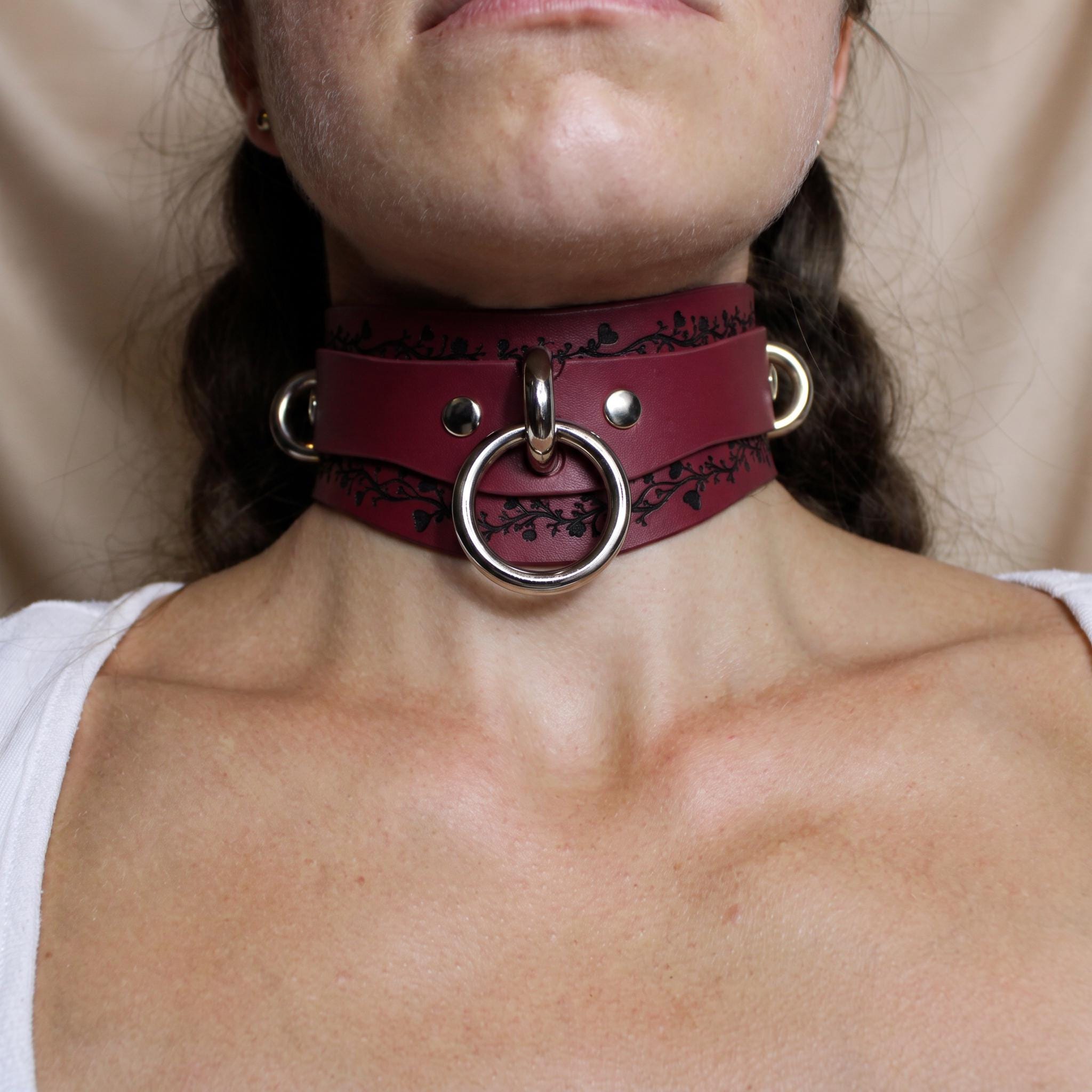 A close-up of a person's neck wearing a burgundy leather choker necklace with a metal ring and two metal rings on either side.