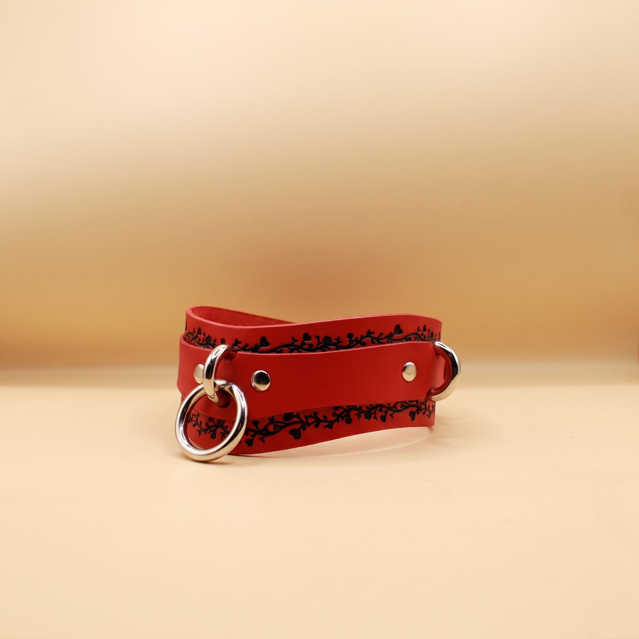 A red leather choker with a metal ring and buckle is shown against a beige background.