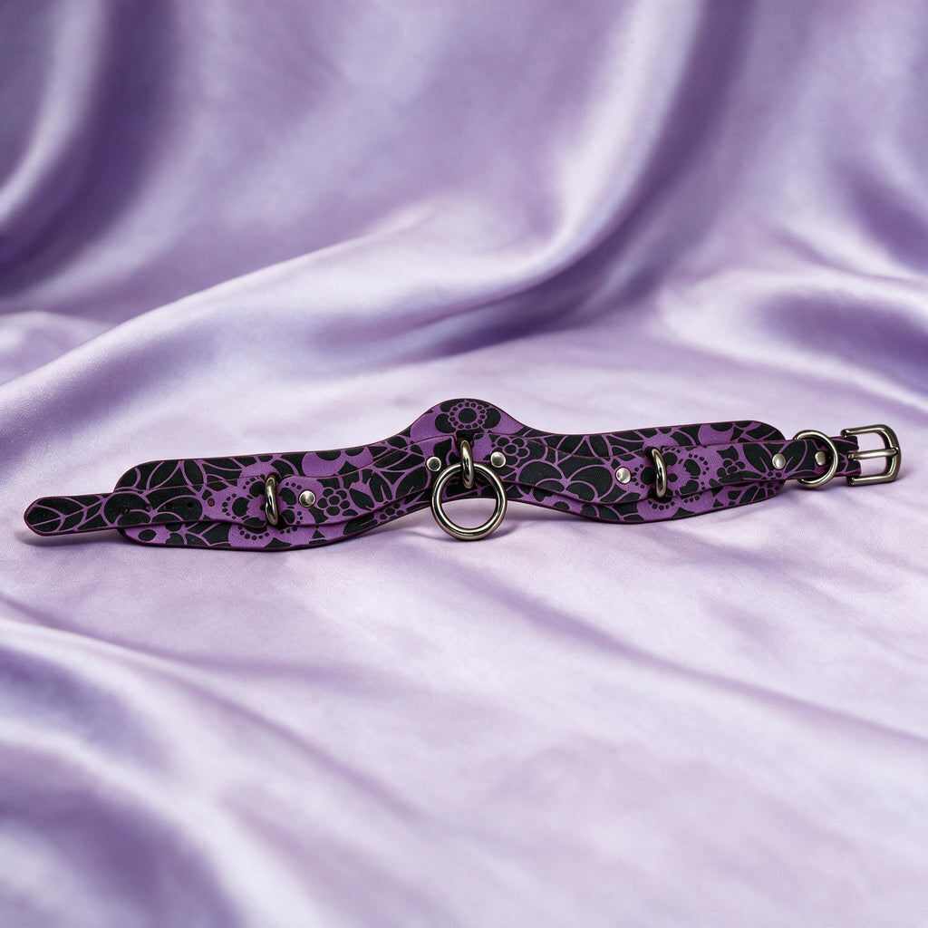 A purple and black patterned choker necklace with metal rings and a buckle is displayed on a purple fabric background.