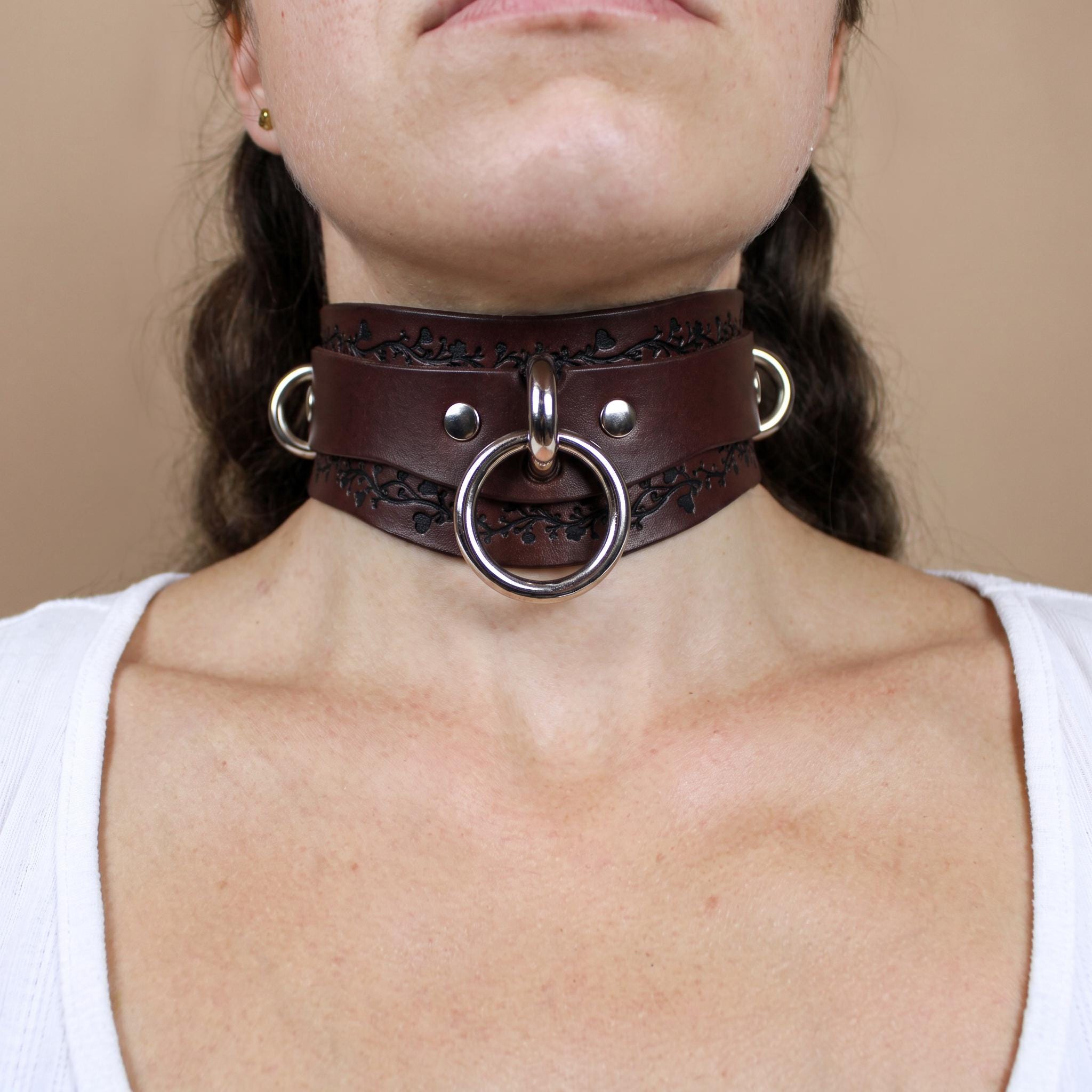 A close-up of a woman's neck wearing a large, ornate leather choker necklace with metal rings and a circular clasp.