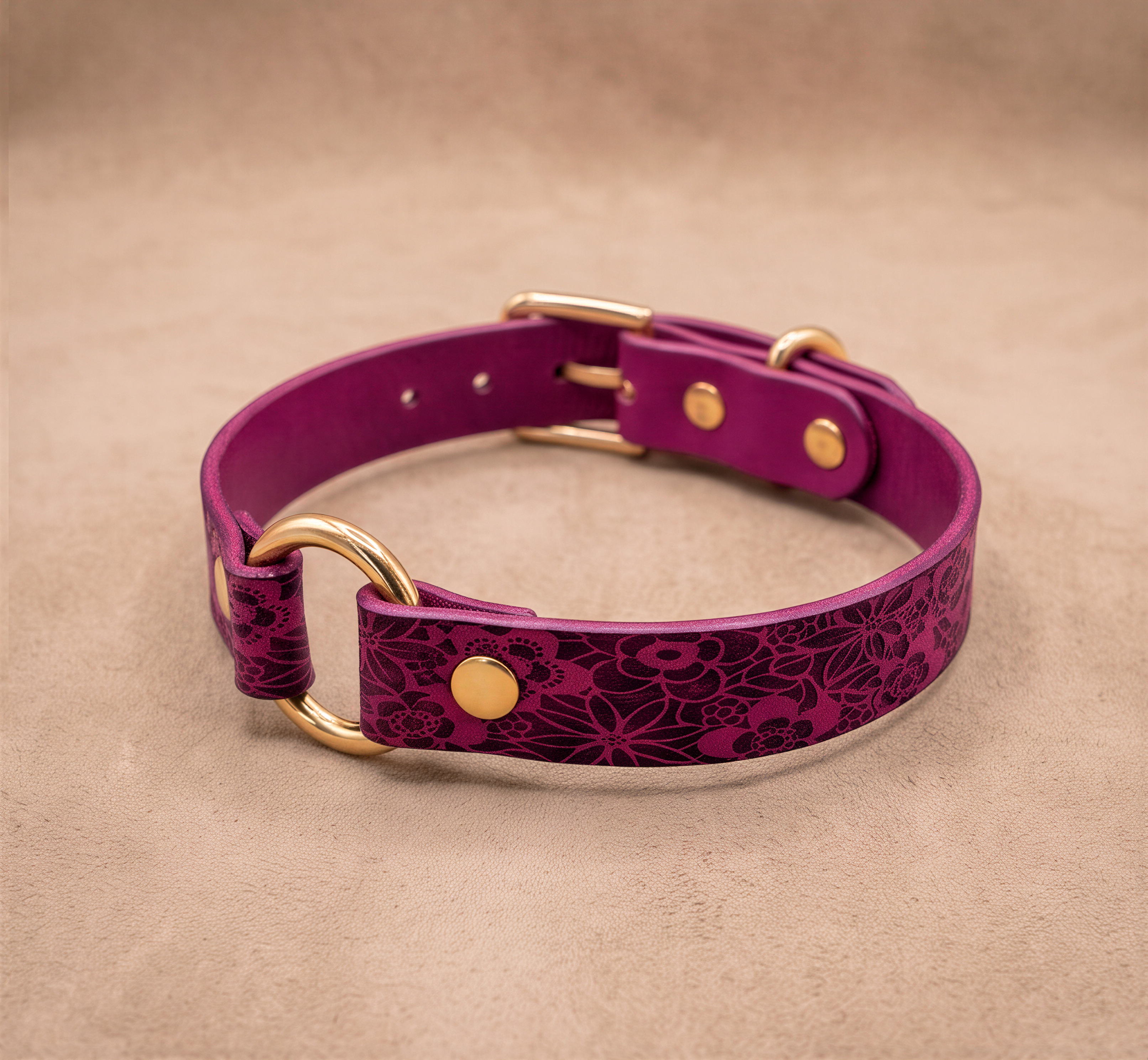A purple leather collar with a floral pattern and gold hardware, including a buckle and ring, is displayed on a beige background.
