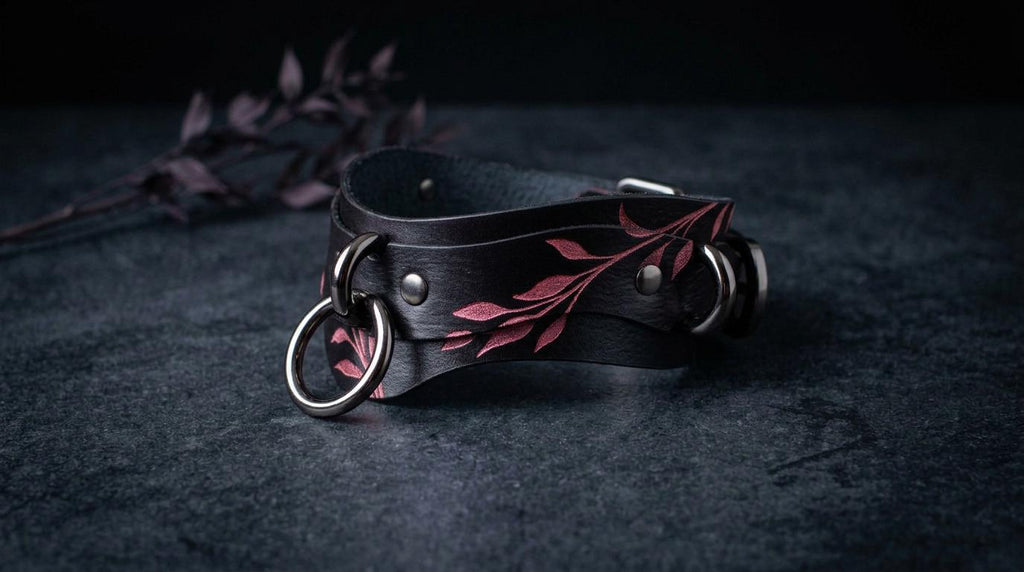 A black leather choker necklace with a leaf design and metal rings is displayed on a dark surface.