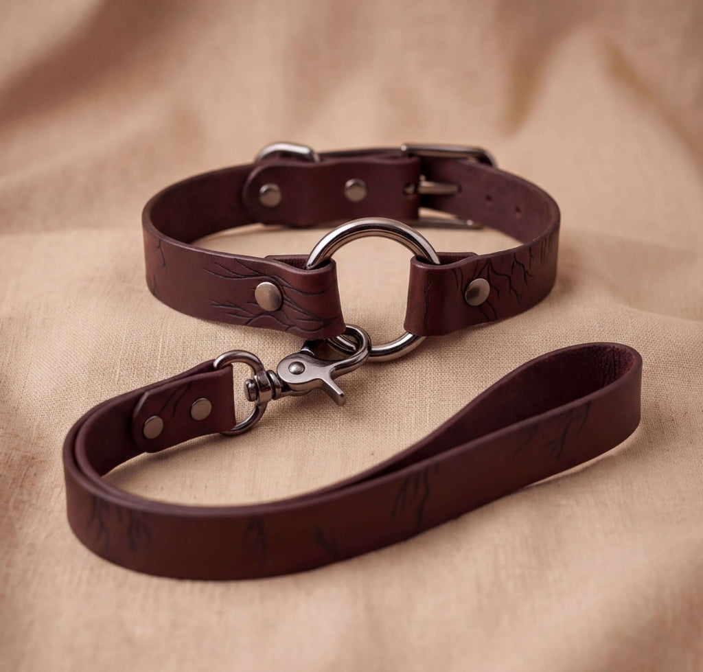 A brown leather dog collar with a metal buckle and a leash attached to it, resting on a beige fabric background.