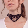 A close-up of a woman's face wearing a black choker necklace with a circular metal ring.