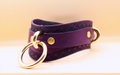A purple leather choker necklace with a gold ring and two gold-colored metal rings on either side.