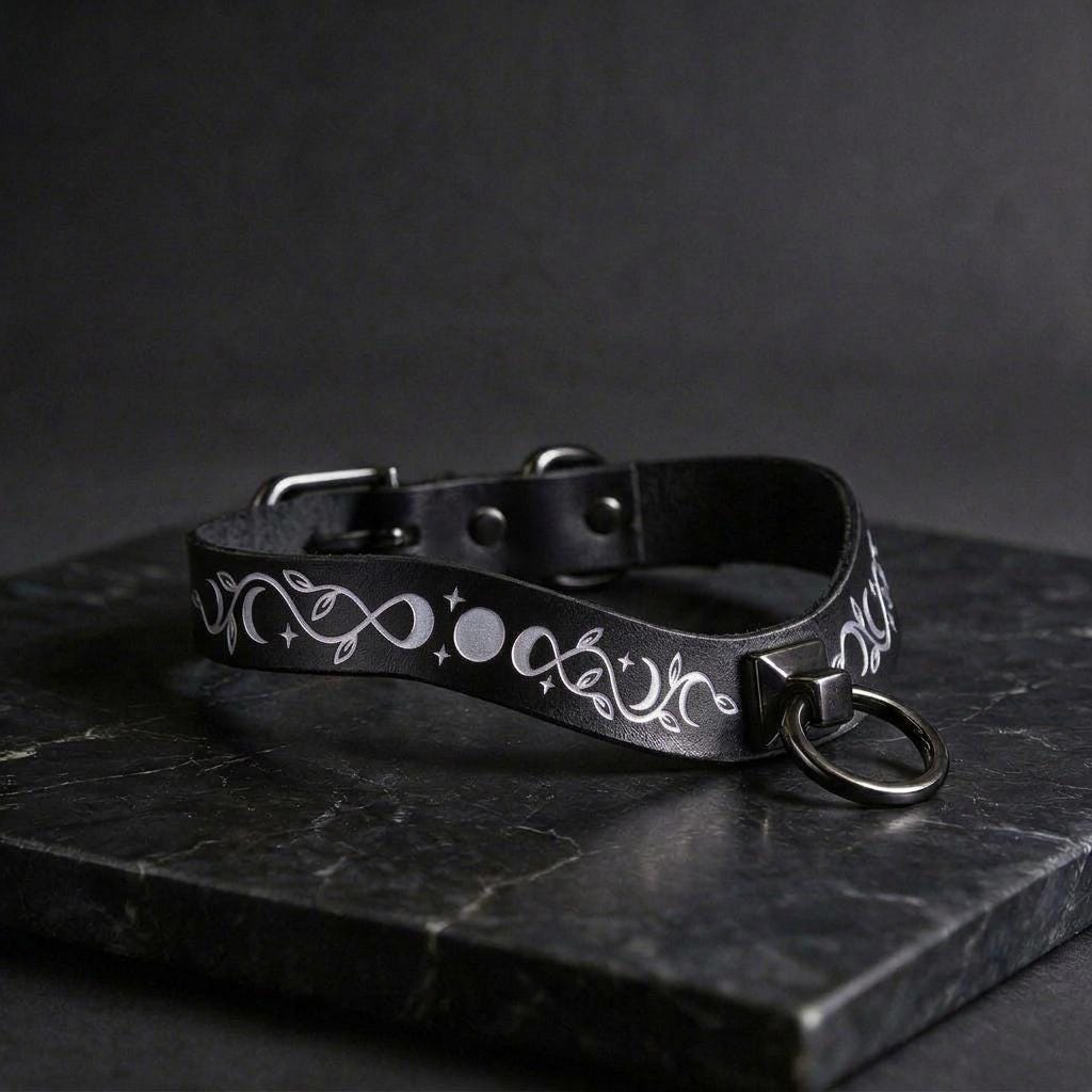A black leather collar with a silver metal ring and decorative white designs on a dark surface.