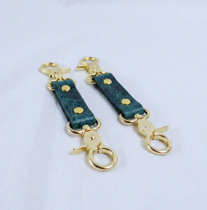 A pair of green leather straps with gold metal clasps and hooks, placed on a white surface.