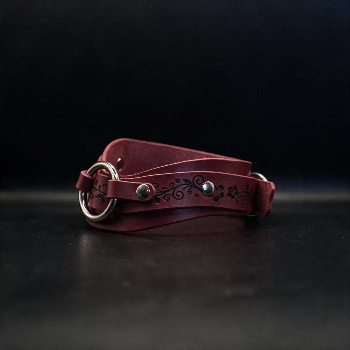 A burgundy leather belt with a floral design on the buckle is displayed against a black background.
