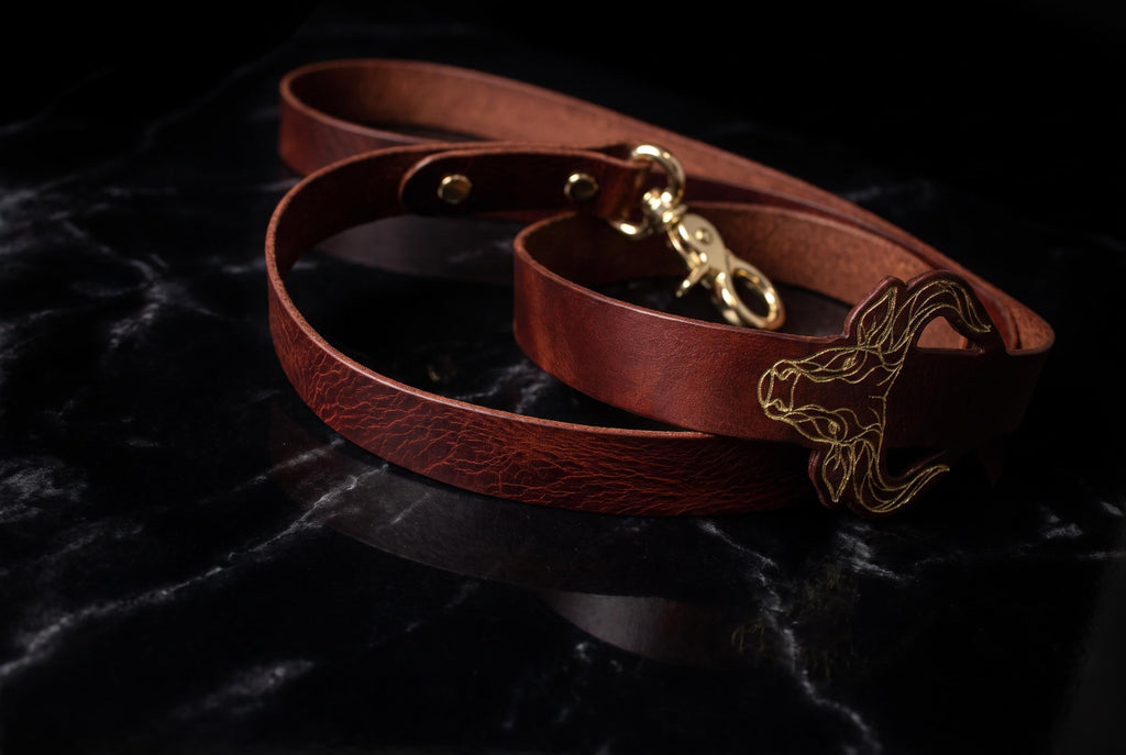 A brown leather leash with a gold buckle and a gold-colored buckle is resting on a black marble surface.
