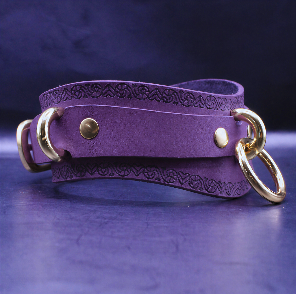 A purple leather strap with gold rings and a decorative pattern on it, resting on a dark surface.