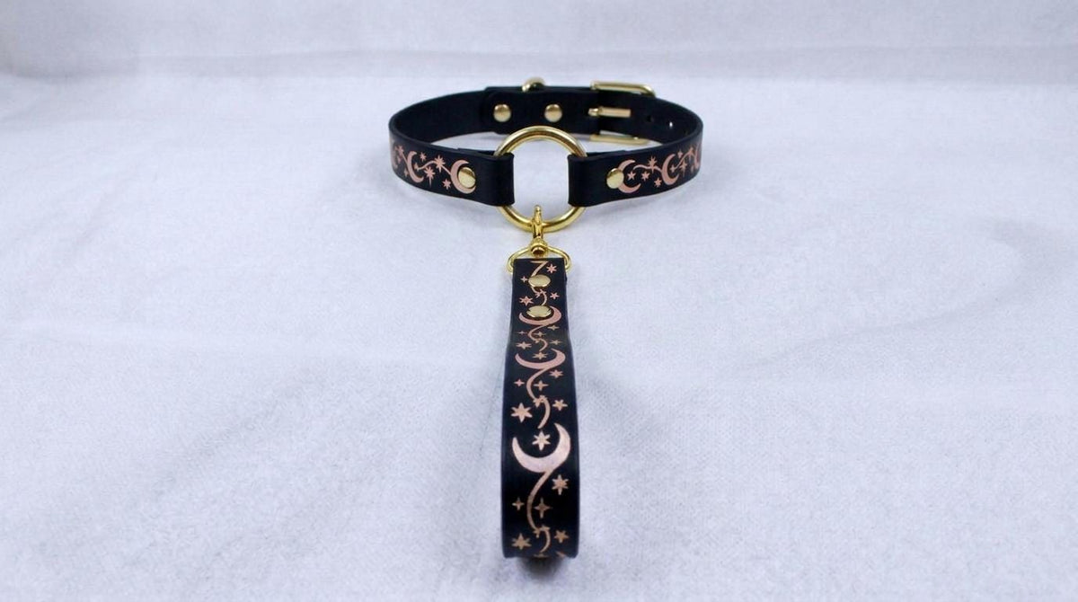 A black collar with a gold-colored buckle and a decorative strap featuring a moon and star pattern.