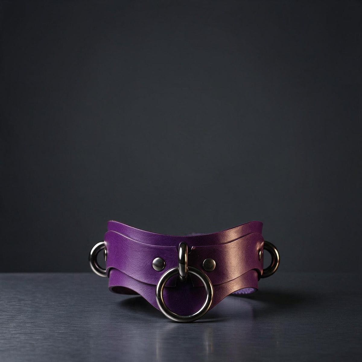 A purple leather choker necklace with metal rings and a circular clasp is displayed against a dark background.