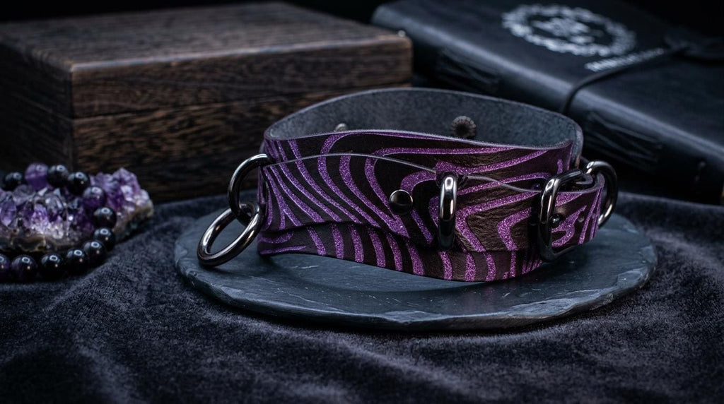 a purple zebra-striped leather choker necklace with metal rings, displayed on a black surface alongside a wooden box and a black book.