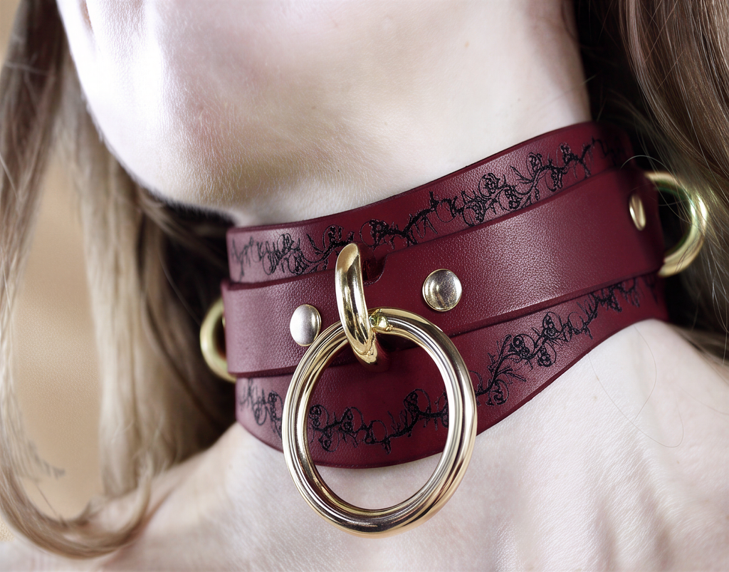 A close-up of a burgundy leather choker necklace with a gold ring and two gold rings on either side.