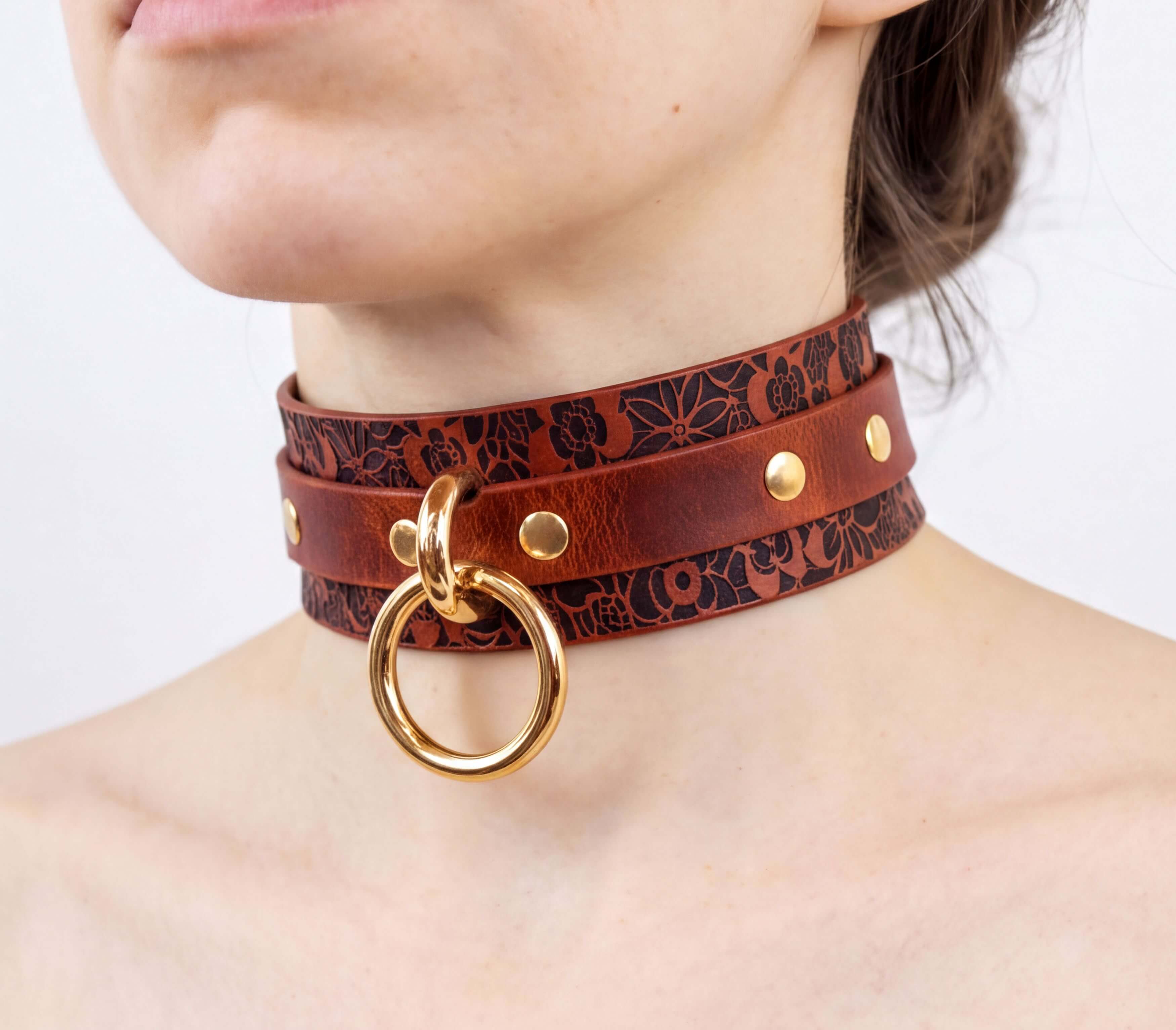 A close-up of a woman's neck wearing a brown leather choker necklace with a gold ring and studs.