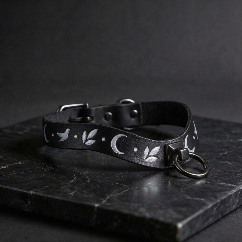 A black leather collar with a silver metal ring and a white floral design on it, resting on a dark surface.