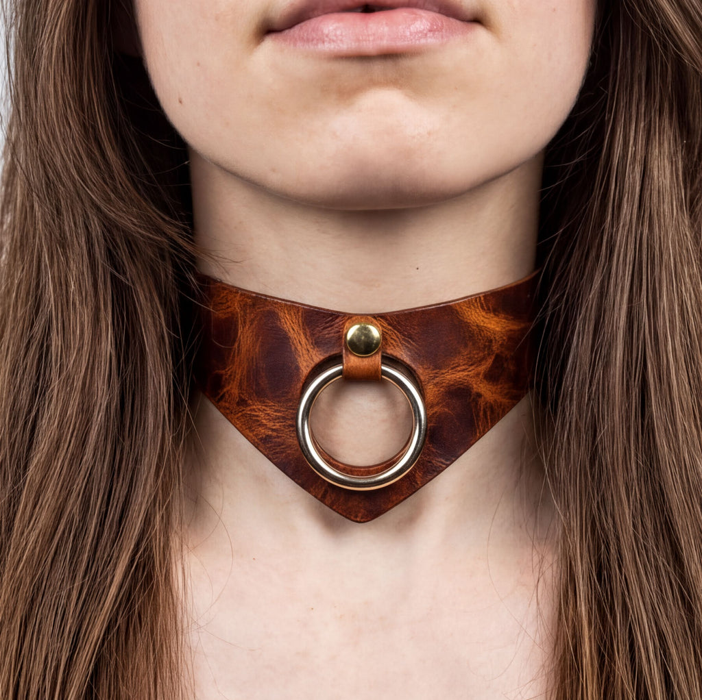 A close-up of a woman's neck and chest area, featuring a leather choker necklace with a metal ring and a gold-colored buckle.