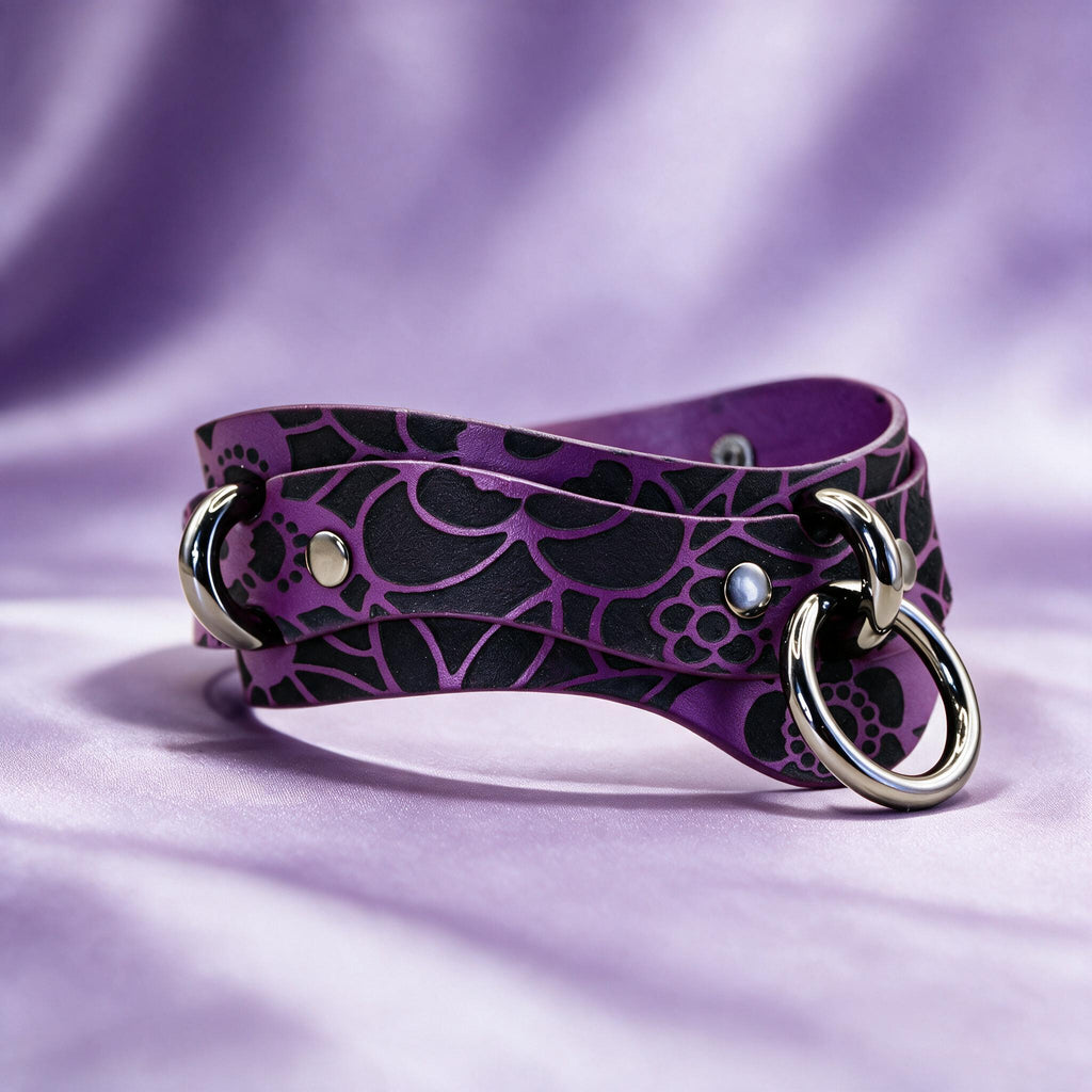 A purple leather choker necklace with a black floral pattern and metal rings on the ends, resting on a purple background.