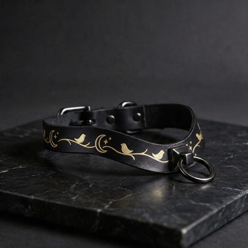 A black leather collar with a gold moon and star design, featuring a metal ring and buckle, is displayed on a black marble surface.