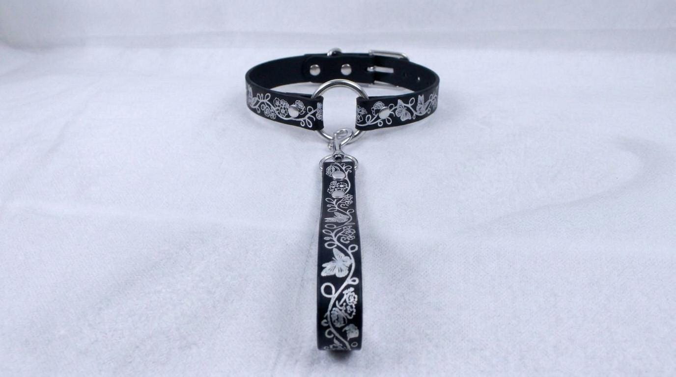 A black collar with a silver buckle and a silver handle attached to it, placed on a white surface.