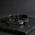 A black leather collar with gold leaf designs and a metal ring on the front, resting on a dark surface.