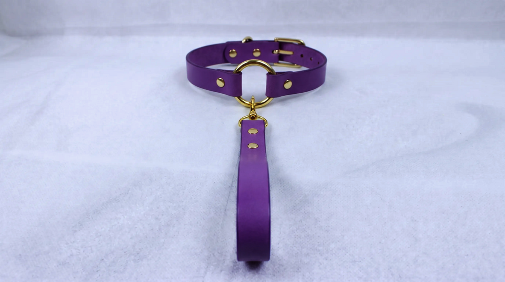 A purple leather collar with a gold-colored buckle and a long strap is displayed on a white surface.
