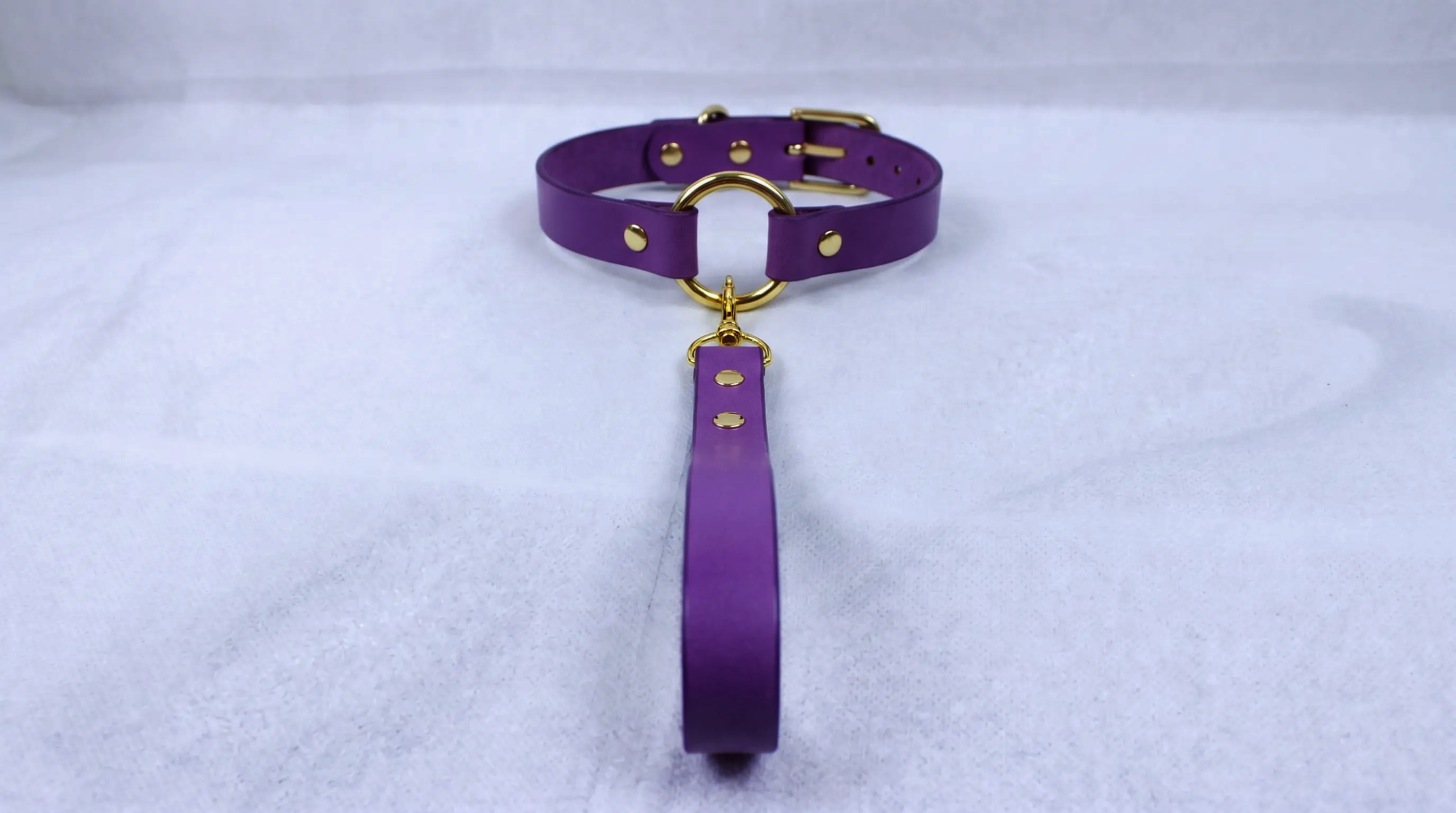 A purple leather collar with a gold-colored buckle and a long strap is displayed on a white surface.