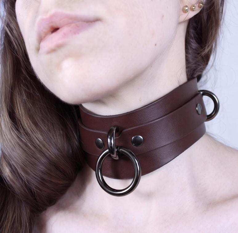 A close-up of a woman's neck wearing a brown leather choker necklace with metal rings.
