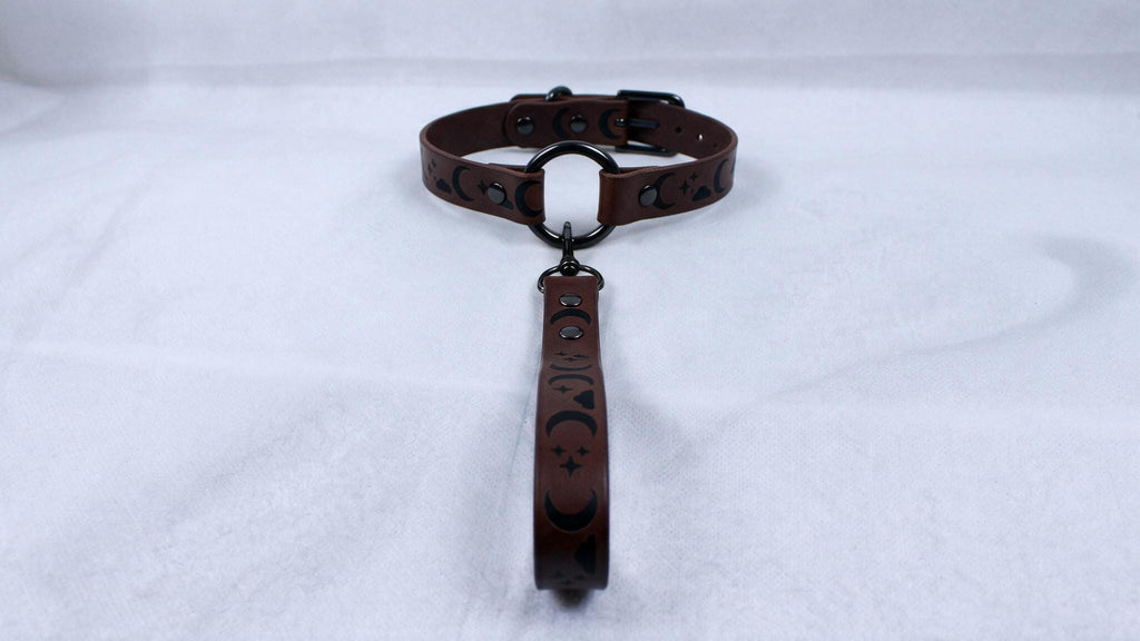 A brown leather collar with a black metal buckle and a black handle attached to it, resting on a white surface.