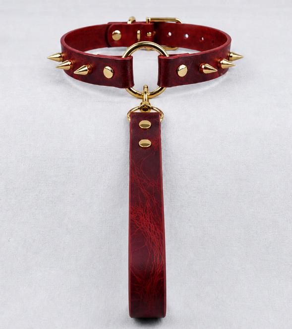 A red leather choker collar with gold studs and a gold-colored loop, hanging from a red leather strap.