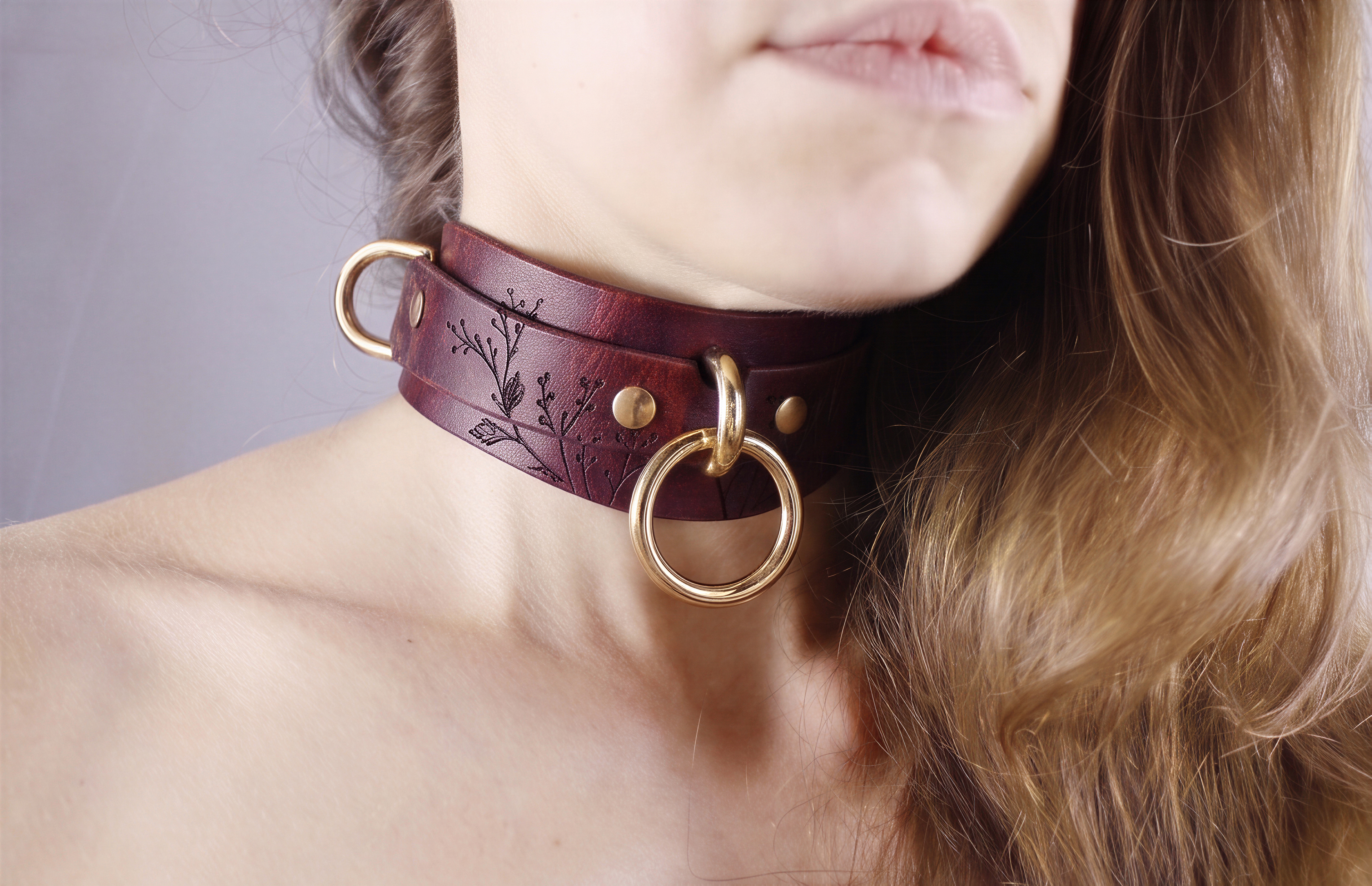 A close-up of a woman's neck and chin wearing a burgundy leather choker necklace with gold rings.